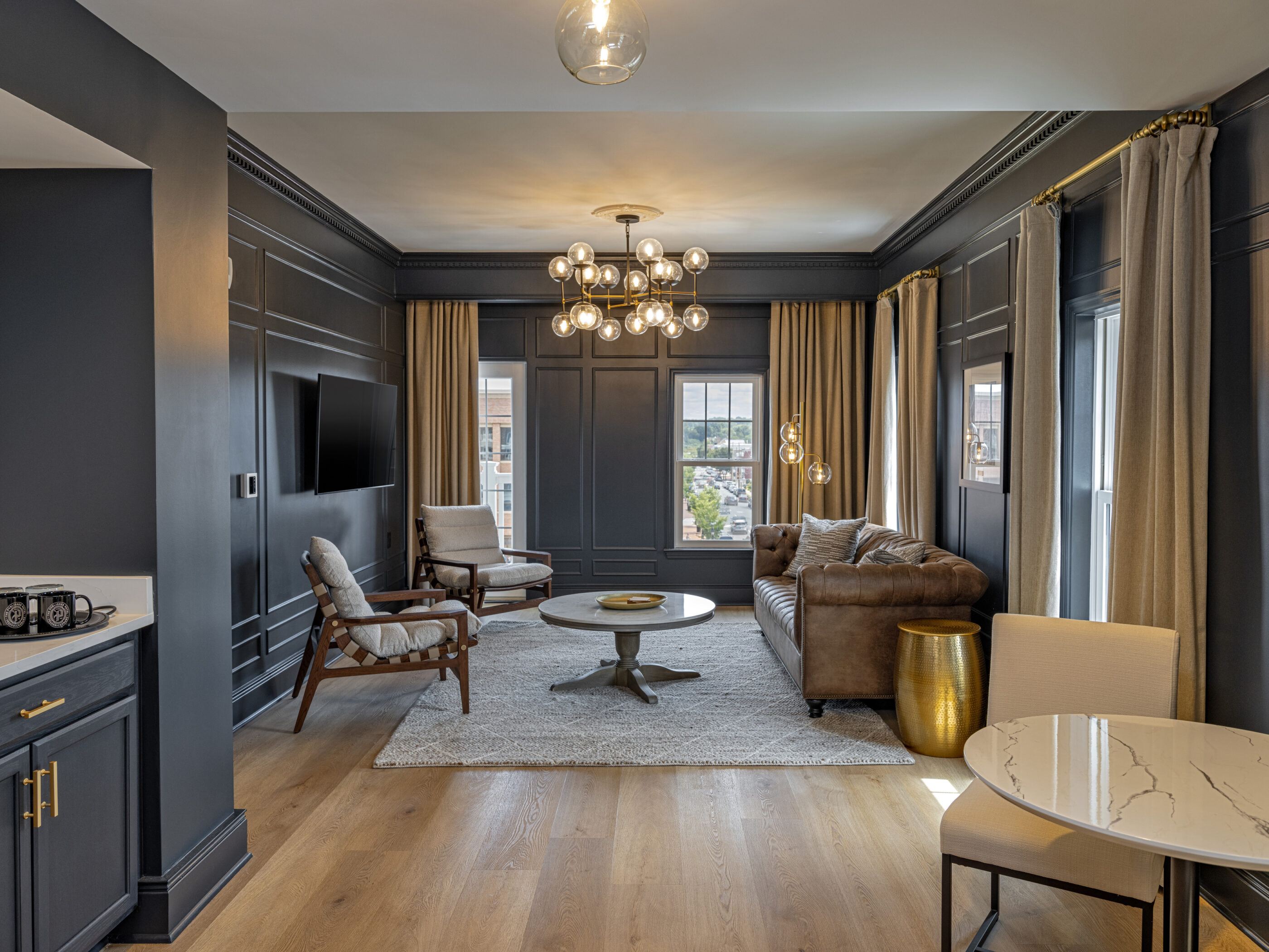 Stylish hotel suite living room with charcoal wainscoting, a tufted leather sofa, modern chandeliers, and a small dining nook.