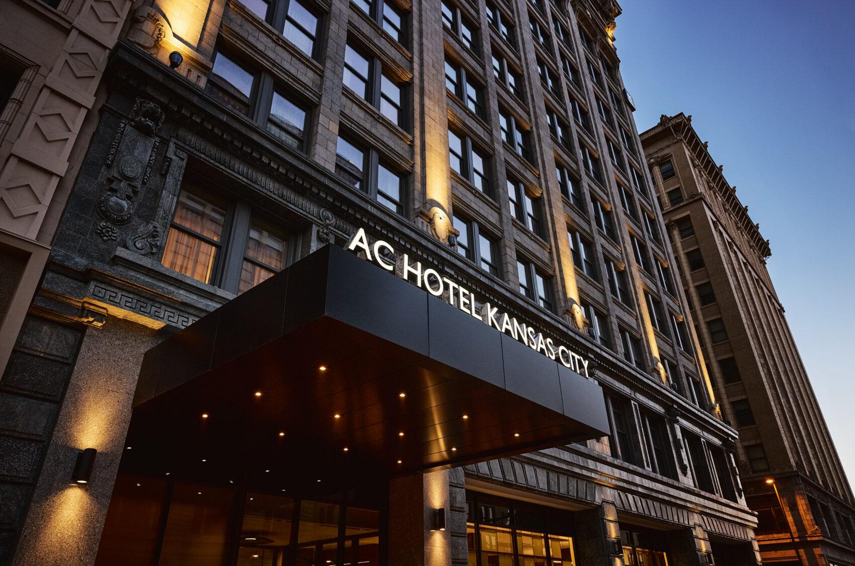 Low-angle exterior view of a historic hotel building featuring a lit canopy entrance that reads "AC HOTEL KANSAS CITY."