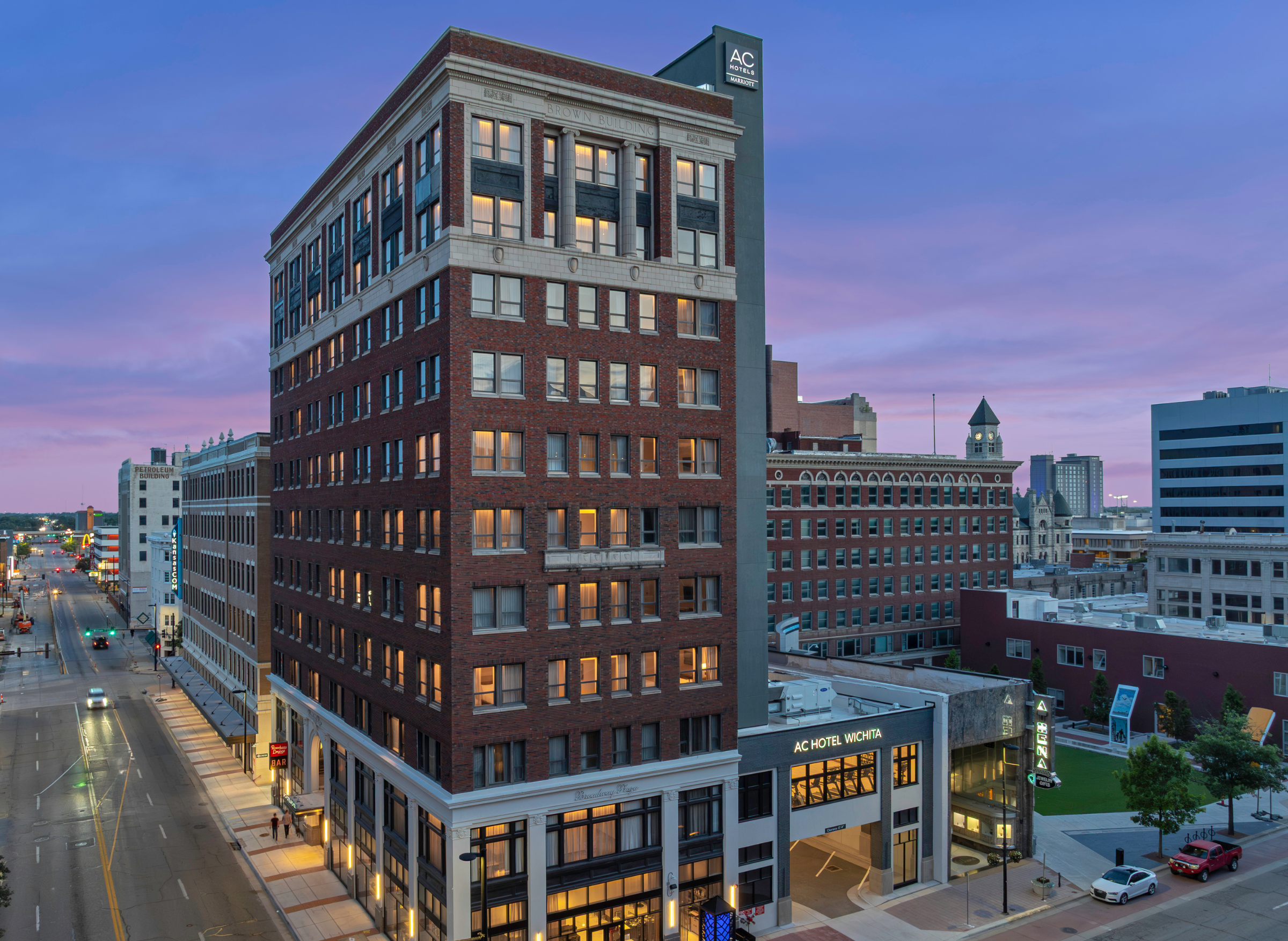 Exterior view of the AC Hotel Wichita building at dusk, featuring a historic brick facade and illuminated city streets below.