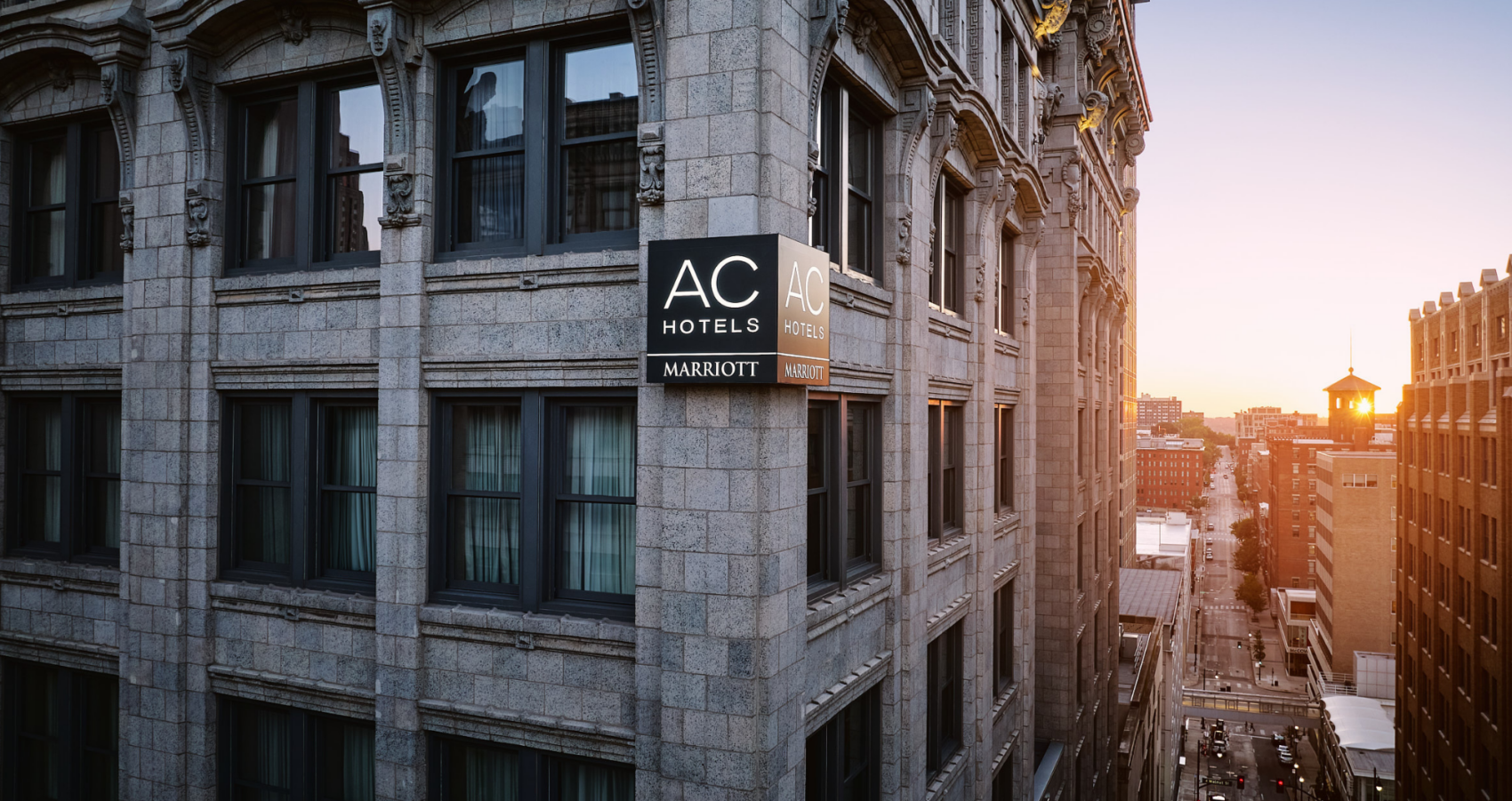 Exterior side angle of the AC Hotel building showing the illuminated signage on its historic stone facade above a sunlit city street at sunset.