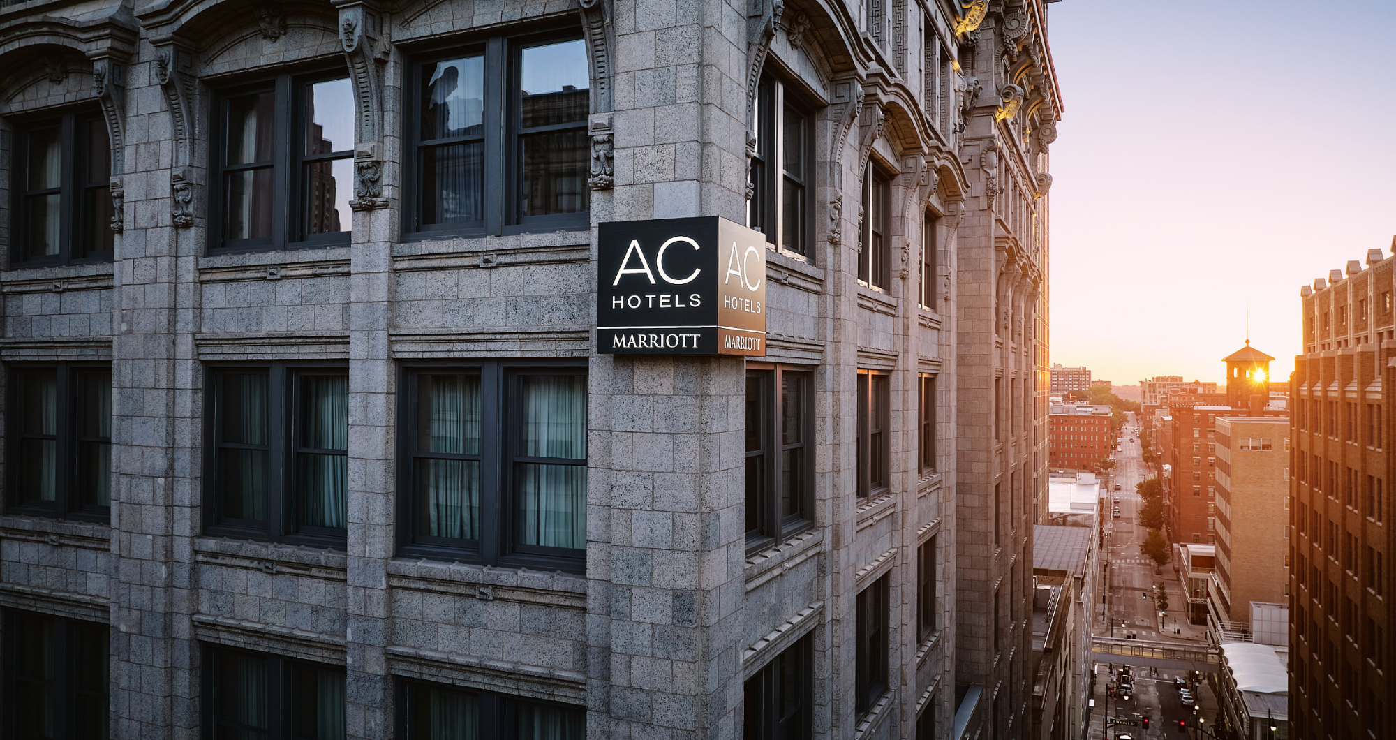 Exterior side angle of the AC Hotel building showing the illuminated signage on its historic stone facade above a sunlit city street at sunset.