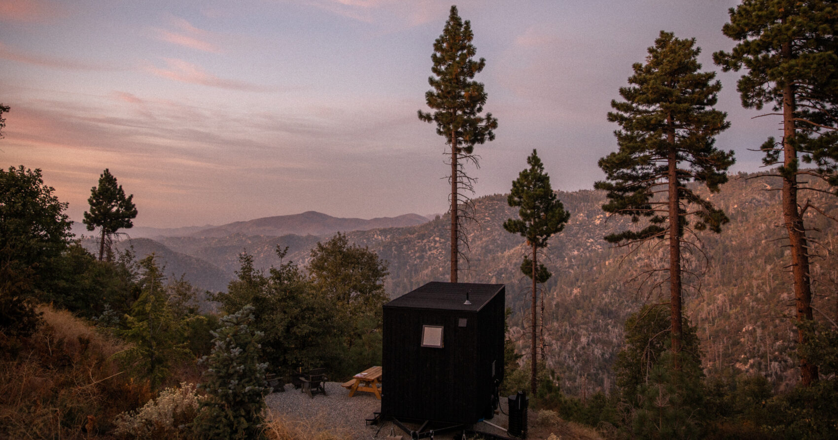 Small black getaway cabin nestled in a wooded forest landscape with mountains in the background under a soft twilight sky.