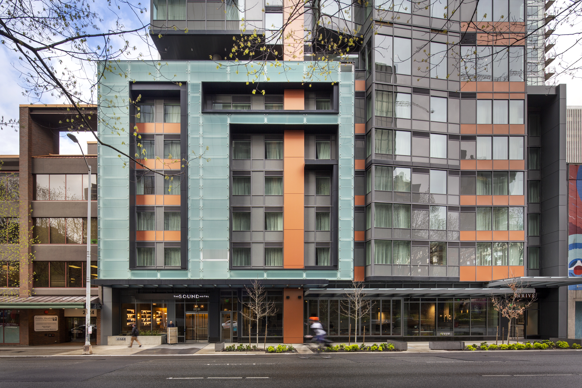 Modern exterior view of The Sound Hotel building featuring a multi-toned facade with glass panels, orange accents, and street-level glass windows.