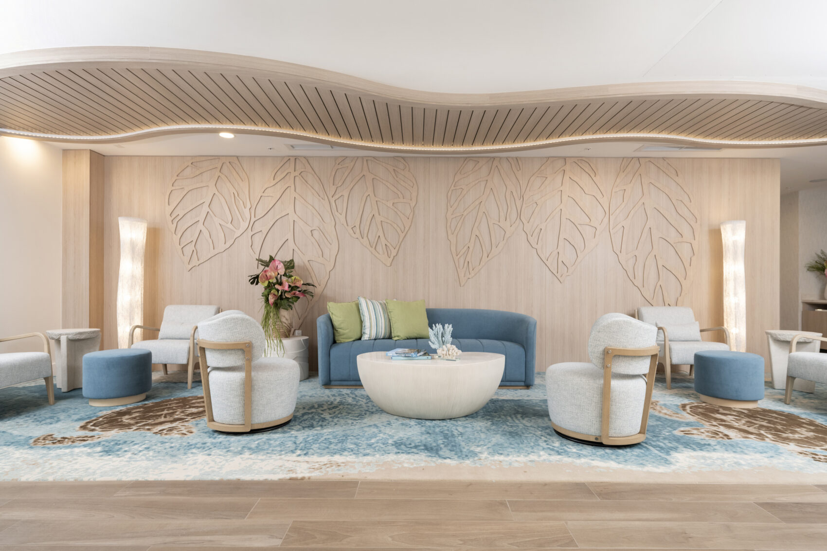 A modern hotel lobby seating area featuring blue and white armchairs, a blue sofa, light wood paneling with sculpted leaf designs, and a wavy slatted ceiling.