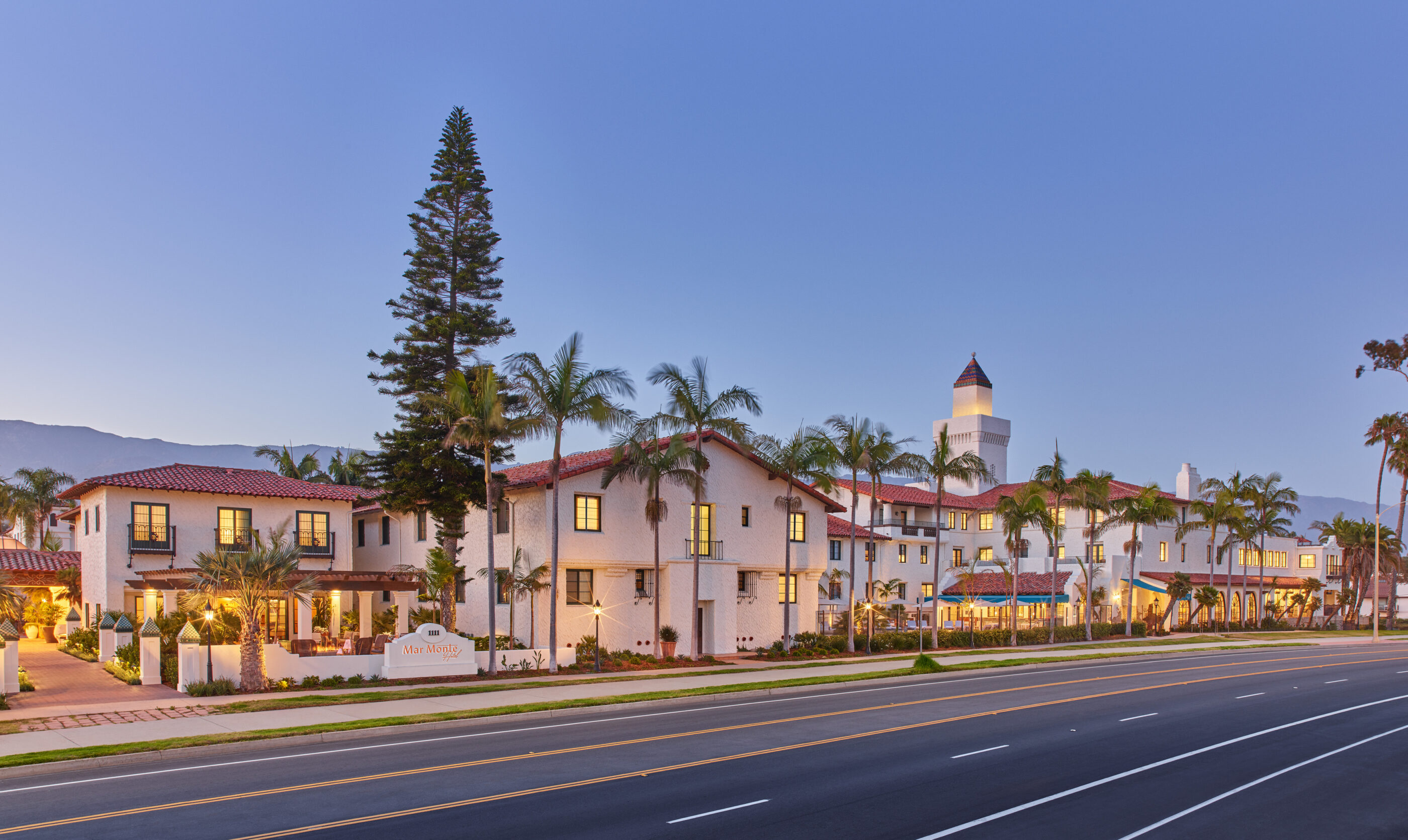 Exterior view of the Mar Monte Hotel property with Spanish Colonial-style architecture, palm trees, and a street in the foreground.