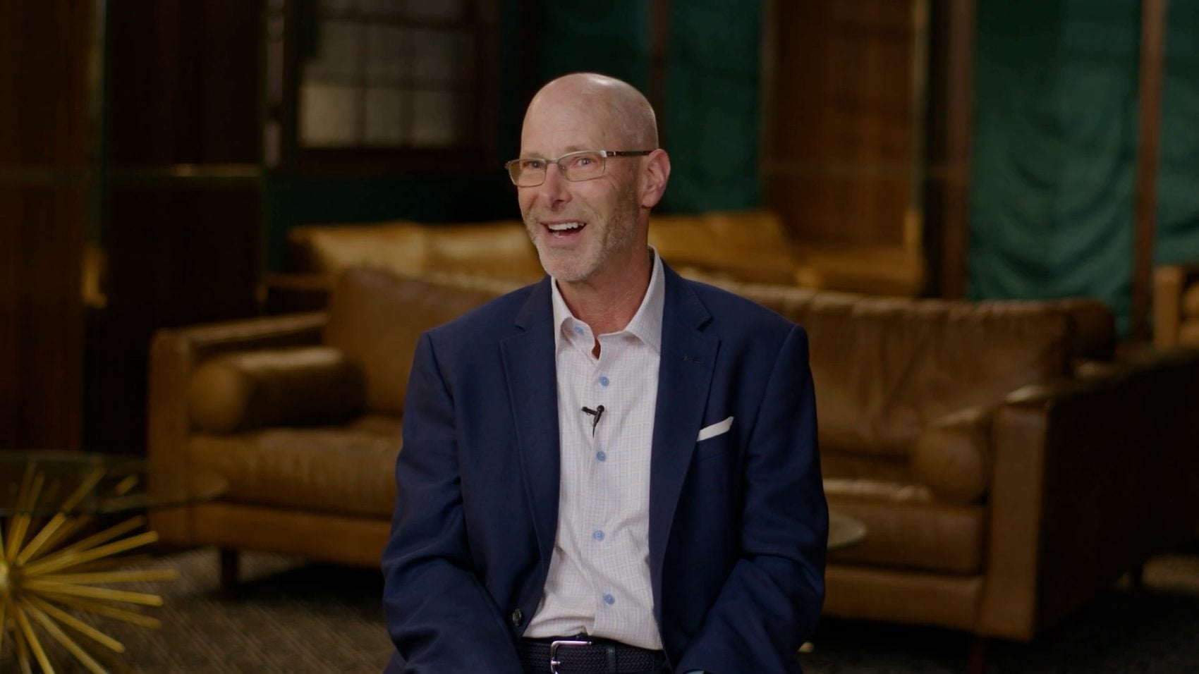 A bald man with glasses and a goatee smiles broadly, wearing a blue suit jacket and a lavalier mic. He sits in a dimly lit room with brown leather couches.