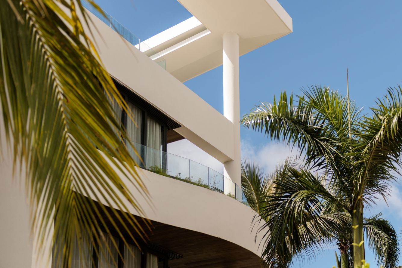 Modern white resort building with curved balconies, glass railings, and palm trees under a sunny sky.