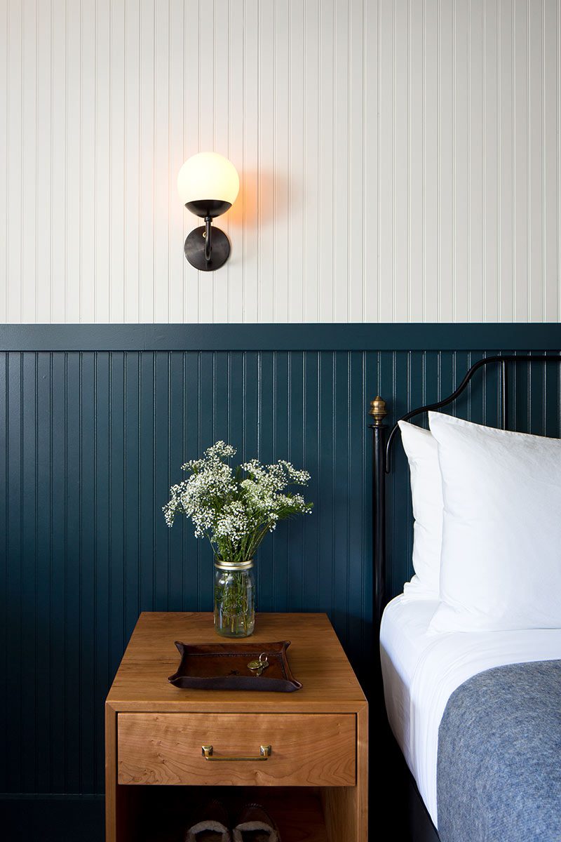 Cozy hotel bedroom setup featuring a wooden nightstand with flowers next to a bed and blue wainscoting wall with a sconce.