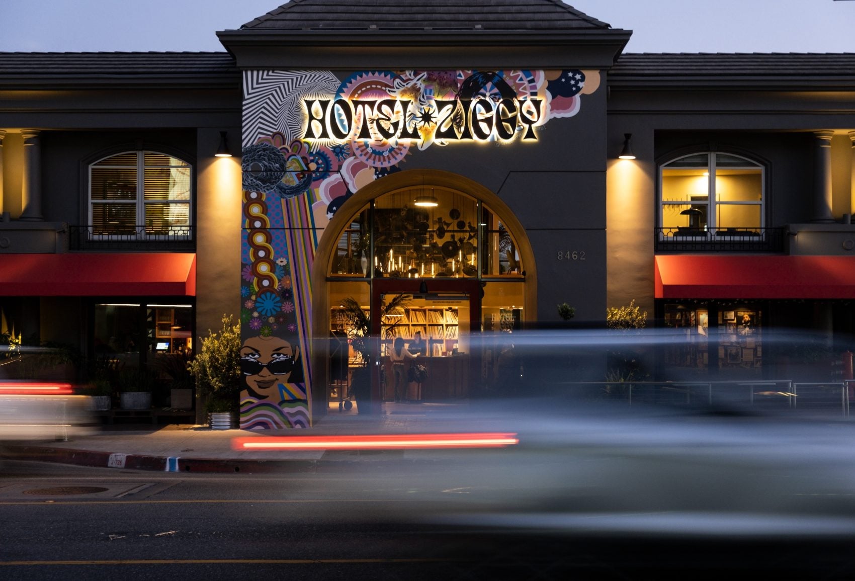Storefront at dusk with a vivid mural covering the facade and a large arched window, warm interior lights visible, and red awnings on either side. Car light trails blur across the street.