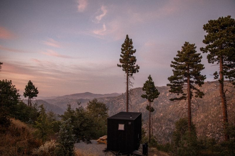 Small black getaway cabin in a forest of tall pine trees overlooking a mountain landscape at dusk at Postcard Cabins.