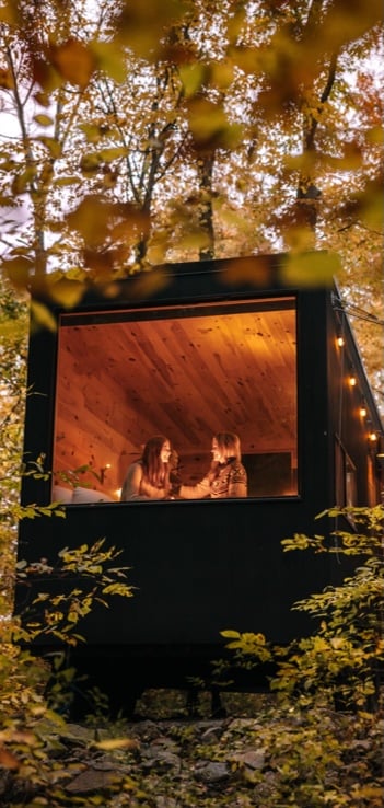 Two women sitting inside a warm, illuminated wooden cabin looking out a large picture window into a lush forest.