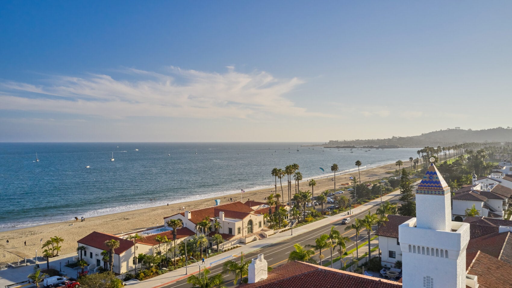 An aerial view of a coastal road and sandy beach lined with palm trees, featuring white Spanish-style buildings with tiled roofs and a prominent white tower with a decorative tiled cap.