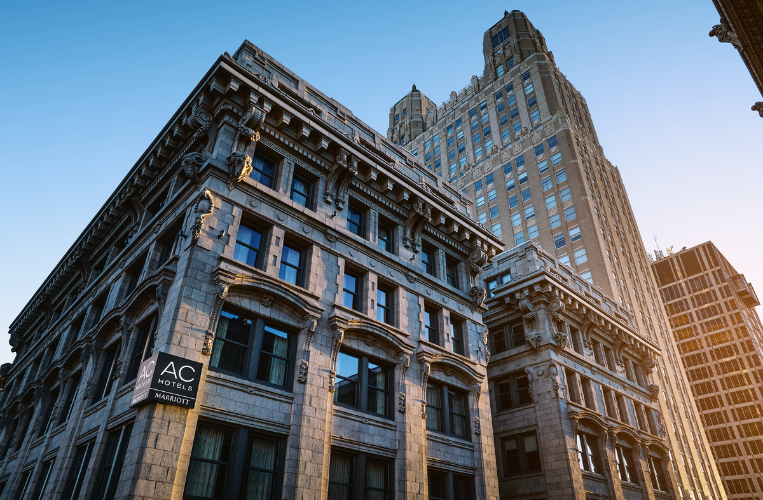 Low-angle shot looking up at a historic multi-story city building with stone facade architecture, large windows, and an "AC Hotels Marriott" sign on the corner.