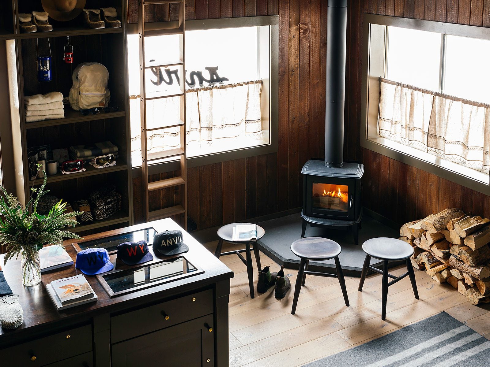 A cozy rustic cabin interior featuring a wood-burning stove with a lit fire, dark wood-paneled walls, large windows, small stools, and a display desk showcasing hats and accessories.