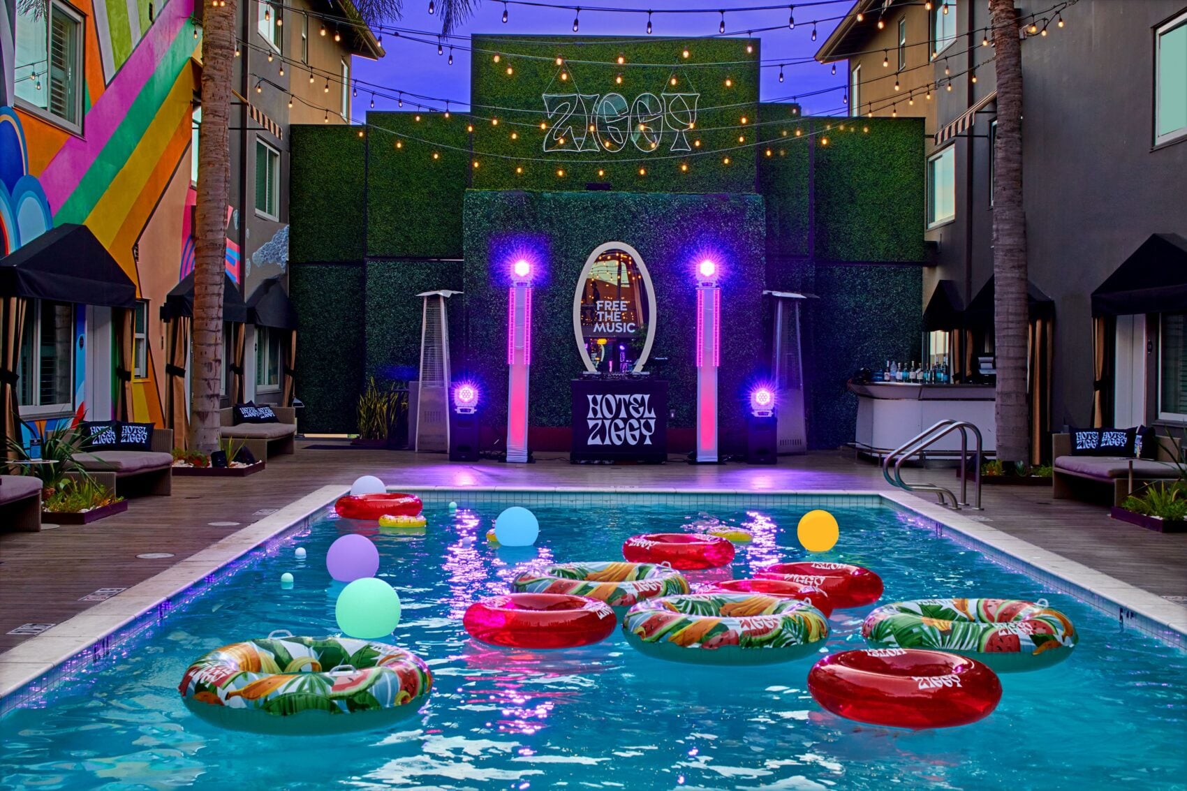 Vibrant outdoor pool area at Hotel Ziggy at dusk, featuring colorful pool floats, purple stage lighting, string lights, and a backdrop banner reading "Free the Music."
