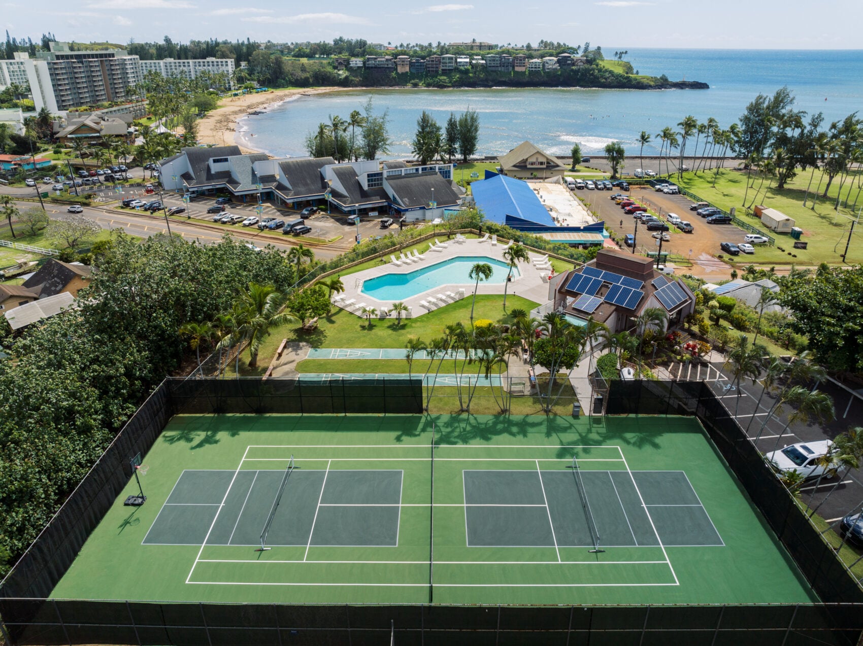 An aerial view of a tropical coastal resort featuring green tennis courts in the foreground, a swimming pool complex, and a scenic bay surrounded by lush foliage and beachfront buildings.