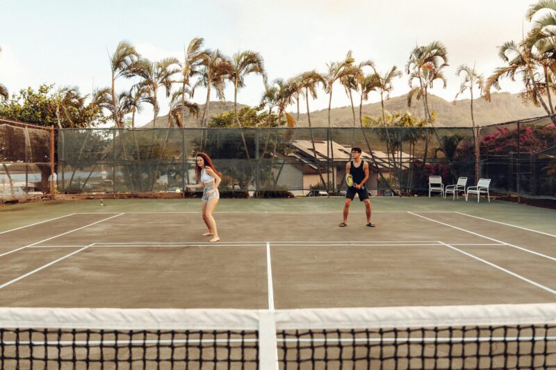 Two people playing tennis on a clay court under palms
