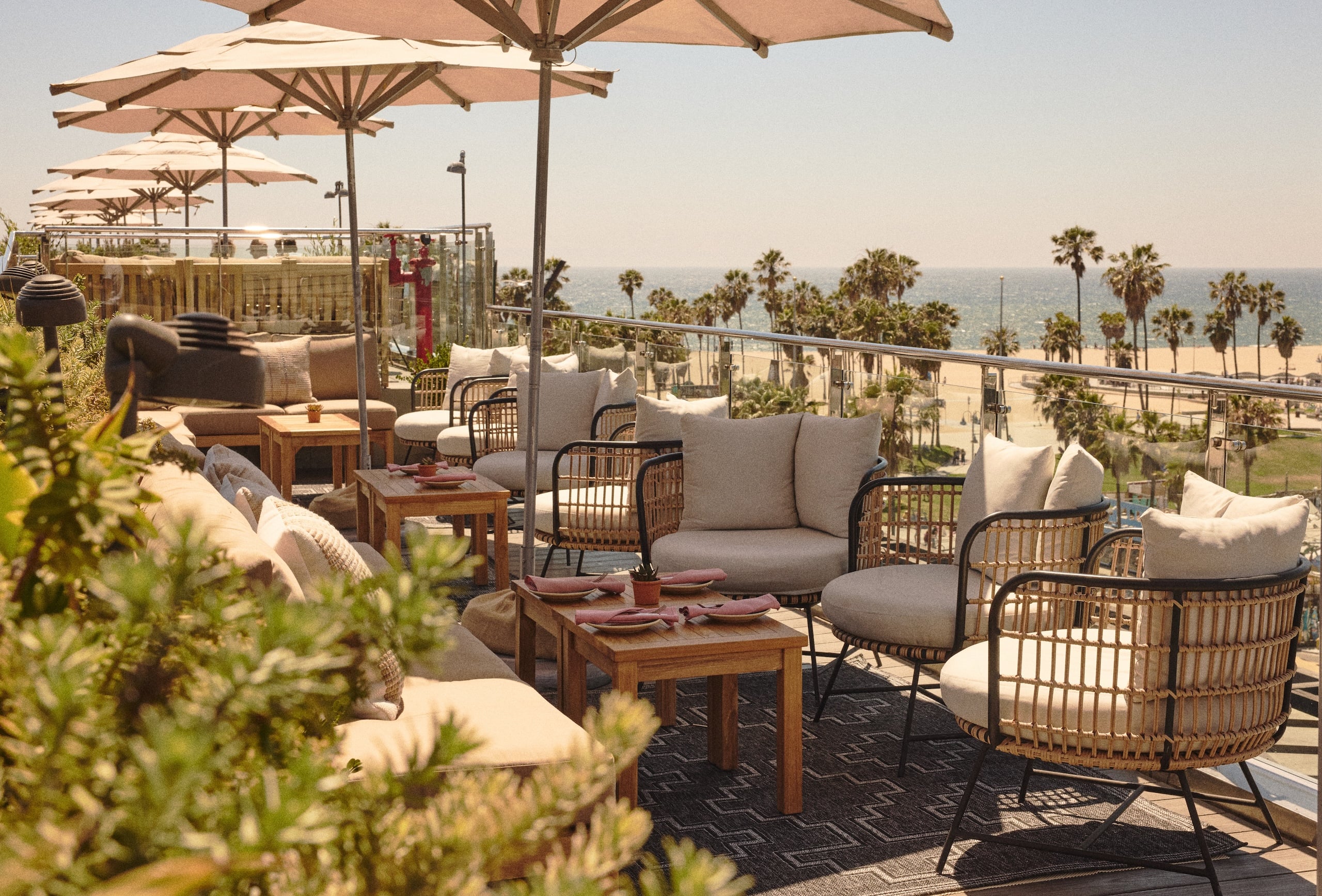 Outdoor beachfront lounge with beige cushioned seating, wicker chairs, wooden tables, and large umbrellas overlooking the ocean.