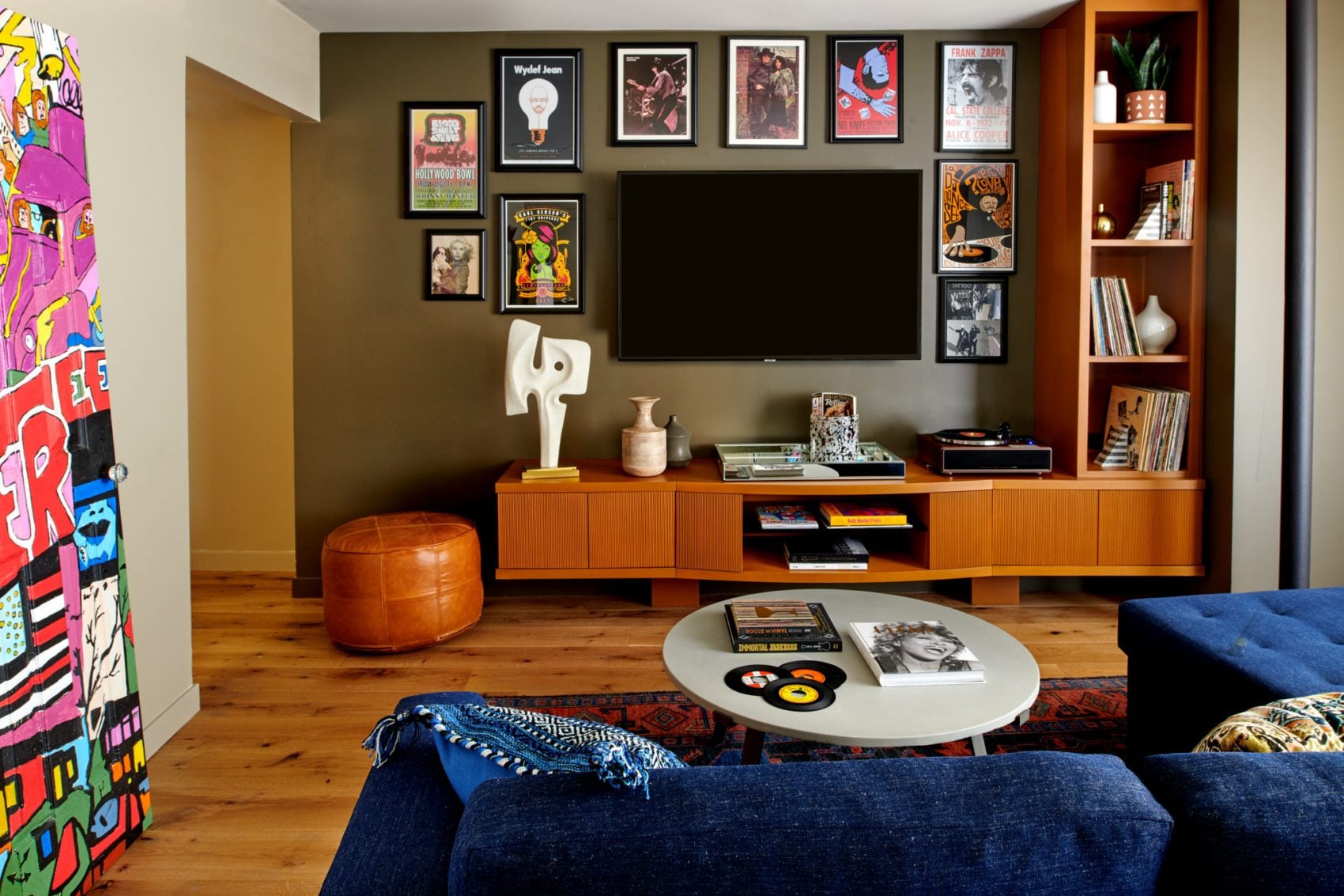 Eclectic hotel lounge with blue sofa, wall-mounted TV, framed posters, and vibrant pop art door.