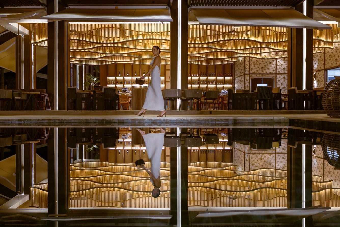 Woman in a white dress walking past a candlelit bar, reflected in a pool