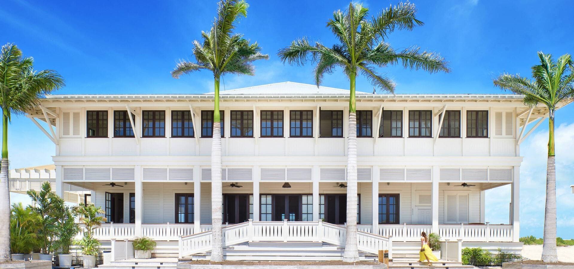 A white two-story building with verandas and dark windows. A woman in yellow walks up steps, flanked by tall palm trees under a clear blue sky.