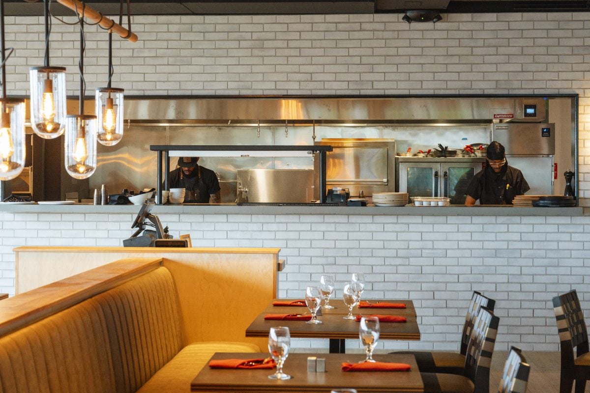 Cooks working in an open restaurant kitchen behind a dining room with set tables