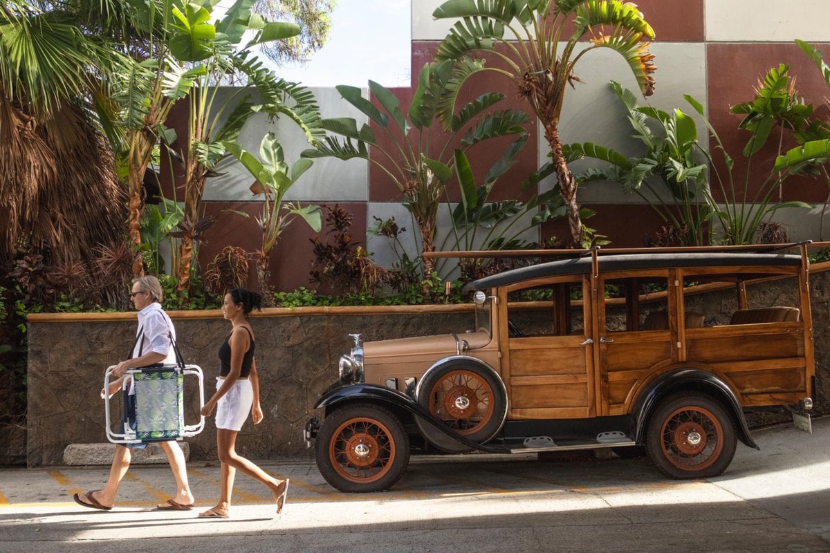 Two guests carrying beach chairs past a vintage wood-paneled car and tropical plants